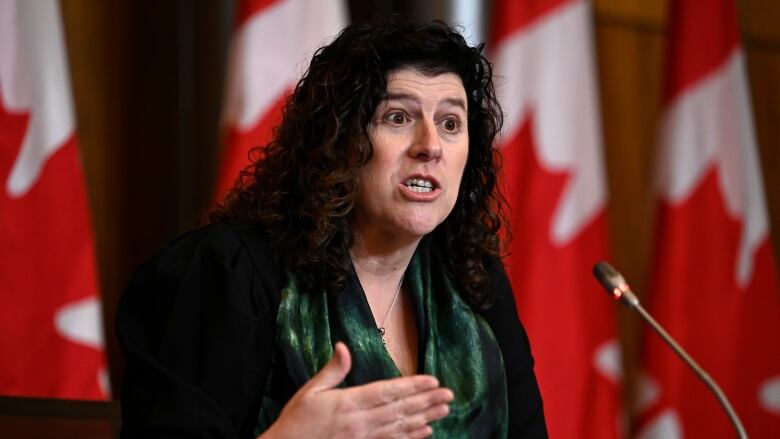 A woman speaks at a microphone in front of a row of Canadian flags.