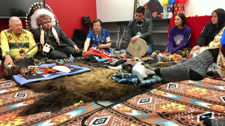 Five people sit on a multi-coloured carpet and buffalo hide rug, including one person holding a drum and one person holding a pipe.