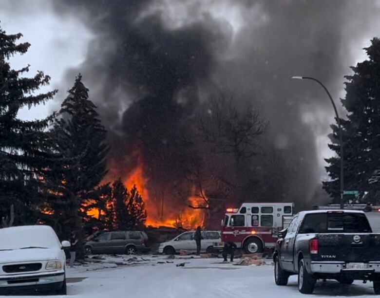 A fire engine and several vehicles surround an area where large flames are burning and sending thick black smoke into the snow-filled air.