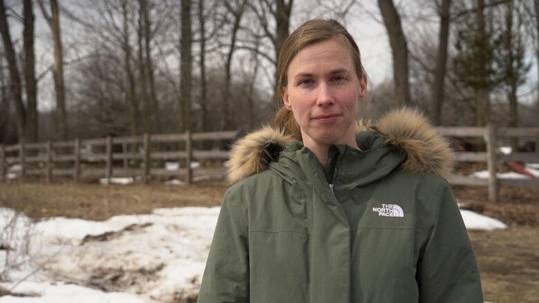 A woman in a jacket stands outside in the snow, looking at the camera. A wood fence is behind her 