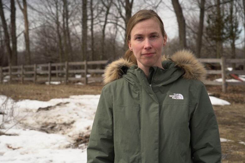 A woman in a jacket stands outside in the snow, looking at the camera. A wood fence is behind her 