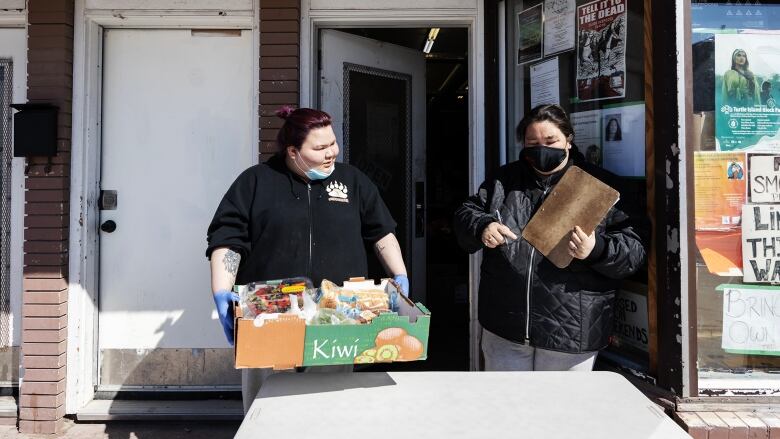 Two women stand behind a table outside a storefront on Selkirk Avenue in Winnipeg's North End. One is holding a clipboard, the other a box of groceries.