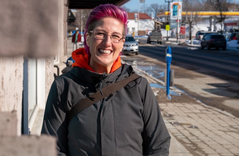 A woman with short pink hair stands on the sidewalk, smiling.