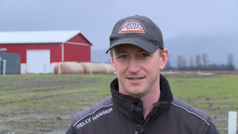 A man wearing a baseball cap stands in front of a barn.