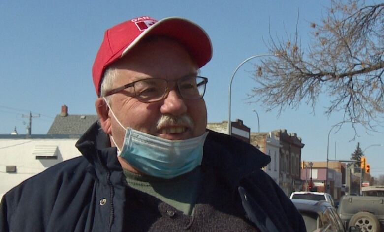 An older man with white hair and a red baseball cap stands on the sidewalk, smiling, his medical mask lowered to his chin.