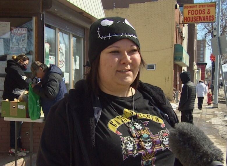 Woman in a black touque and rock'n'roll t-shirt stand in front of the Bear Clan food bank with other small storefronts in the background.