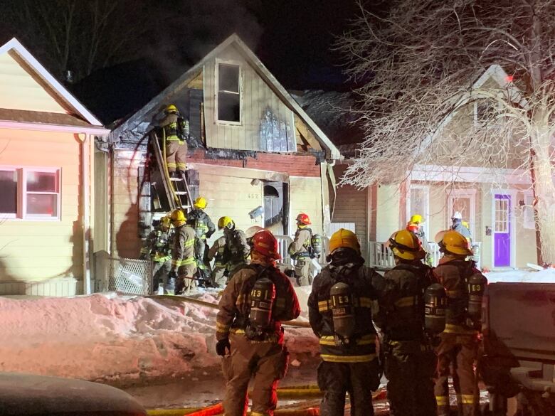 Multiple firefighters stand at a ladder on a fire-damaged house as other firefighters watch from a distance.