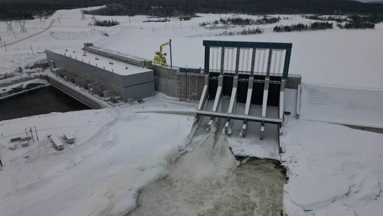 An aerial view of a hydroelectric dam in winter. Water rushes through an open spillway gate.