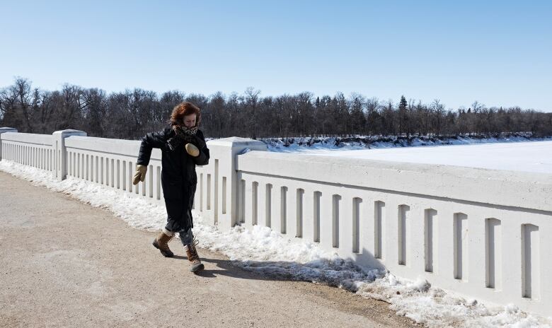 A woman walks quickly across a snowy bridge over a snowy river. Her arms are swinging and face covered in a scarf.