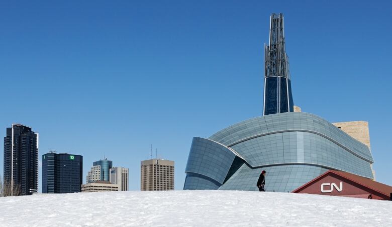 A person is seen in the distance walking across a snow-covered hill. The city skyline is seen in the background.