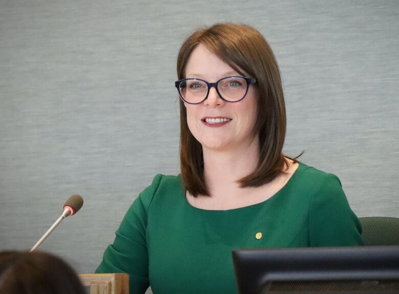 A woman in a green dress and glasses smiles and speaks into a microphone.