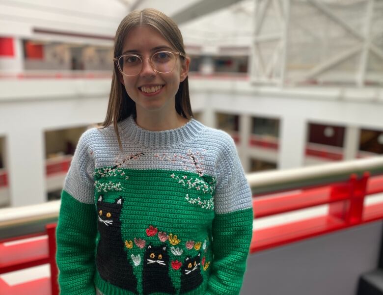 A woman wearing a sweater that depicts a Maud Lewis painting, three black cats on a green background.