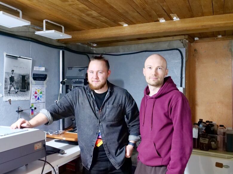 Two men stand next to each other in a room filled with photo processing equipment. 