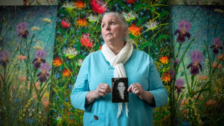 A white woman with long grey hair pulled back in a ponytail holds a black-and-white photo of a teenage girl in front of her chest. The woman is wearing a robin's egg blue cardigan and a white scarf and standing in front of three large, vibrant paintings of flowers.