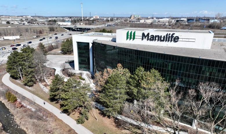 A drone shot of a building with the Manulife logo on the top. It's beside a highway and behind some trees in the foreground. 