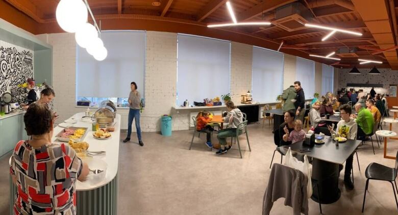 A group of people, including children, are show in a lunchroom. A large banquet at the front holds trays of food and cafe tables and chairs are scattered throughout.