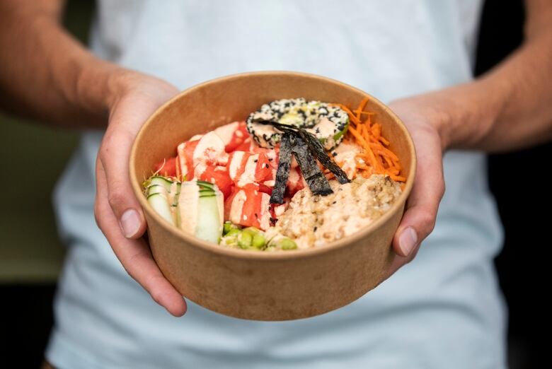 A pair of hands hold a cardboard bowl containing rice, vegetables and sauce.