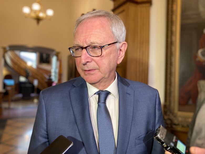 A man stands in the halls of legislature looking at a reporter out of frame. He is wearing a blue suit, white shirt and blue tie, as well as glasses.