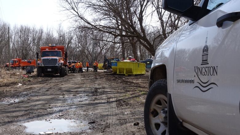 A Kingston logo on the door of a white truck is shown in the foreground. In the background an orange dump truck, green dumpster and workers can be seen at the far end of a muddy road.