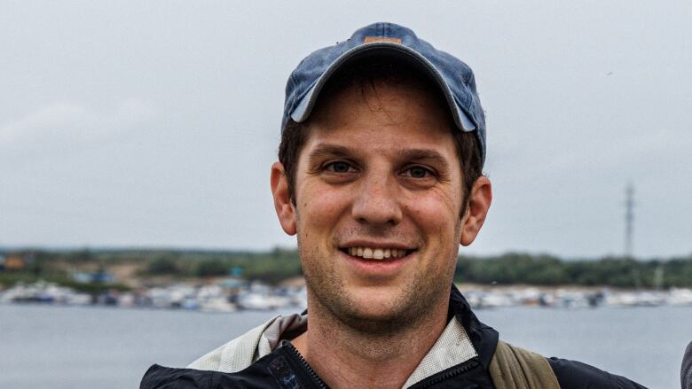 A man in a baseball cap and jacket with backpack straps is shown posing for a photo outdoors.