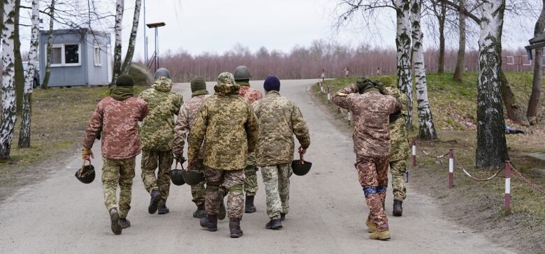 A group of eight men in mismatched military clothing and carrying helmets walks away from the camera down a country road.