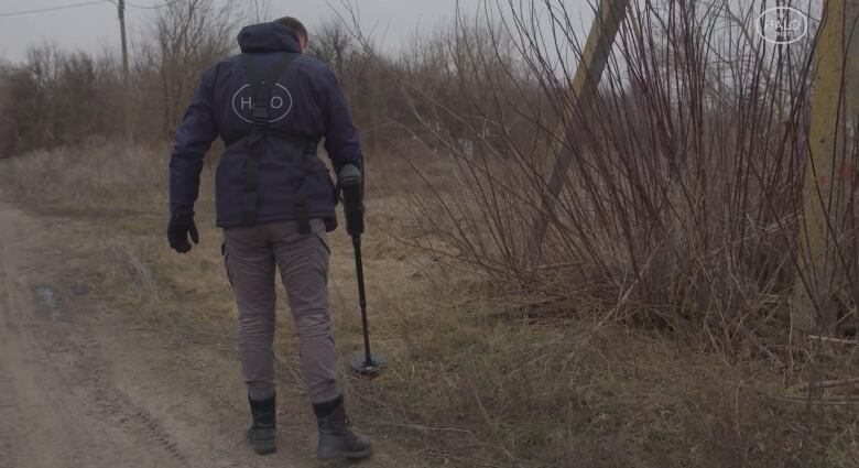 A man in a blue parka uses a metal detector to scan the ground in an forested area alongside a dirt road.