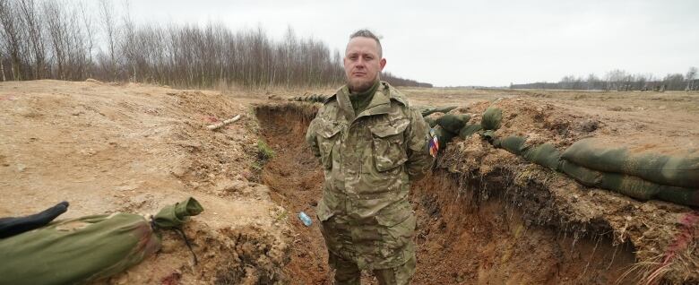 A man in military camouflage clothing stand with his arms behind his back in a dirt trench.