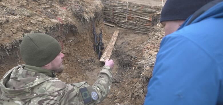 A man in a blue parka watches as another man in Canadian military camouflage clothing gestures at a pair of rifles lying at the bottom of a dirt trench.