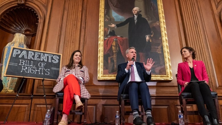 Two women and a man sit in a government building with a painting of George Washington behind them. A chalkboard sign next to the left-most woman reads 'Parents Bill of Rights.'