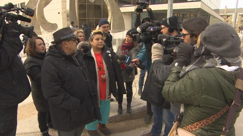 Journalists with microphones and cameras surround a small group of people outside of a law court building.