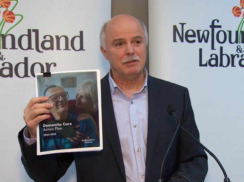 A man wearing a suit stands at a microphone smiling, holding a copy of a report. The cover page reads 'Dementia Care Action Plan 2023-2026, and features an elderly couple smiling at each other.