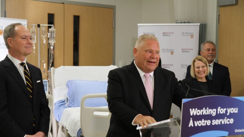 A person in a suit talks and smiles at at a podium in front of a hospital bed and three other people.