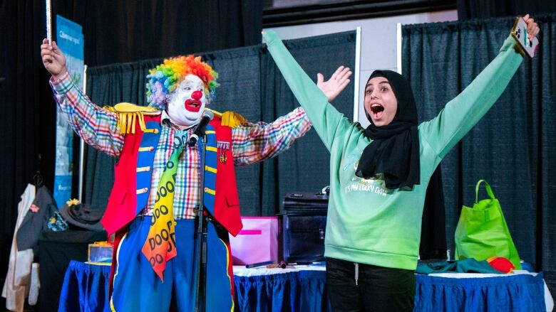 A clown and a girl in a hijab raise their arms in celebration.