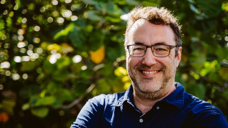 Headshot of Daniel Flander smiling, wearing a blue collared shirt, with greenery in the background. 