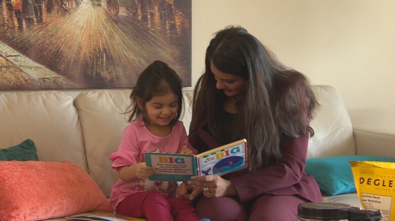 A woman and her young daughter sit on a couch in a living room both holding the same book.