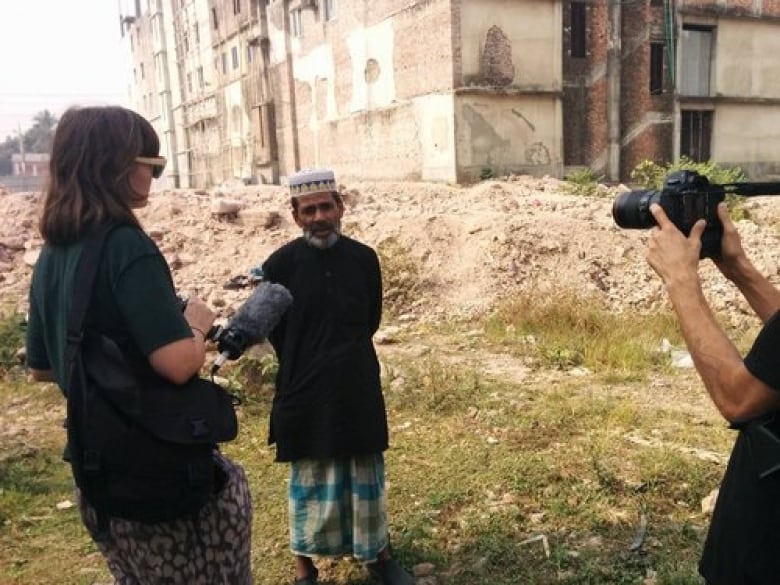 A woman with a camera and microphone speaking to a Bangladeshi man backdropped by rubble. 