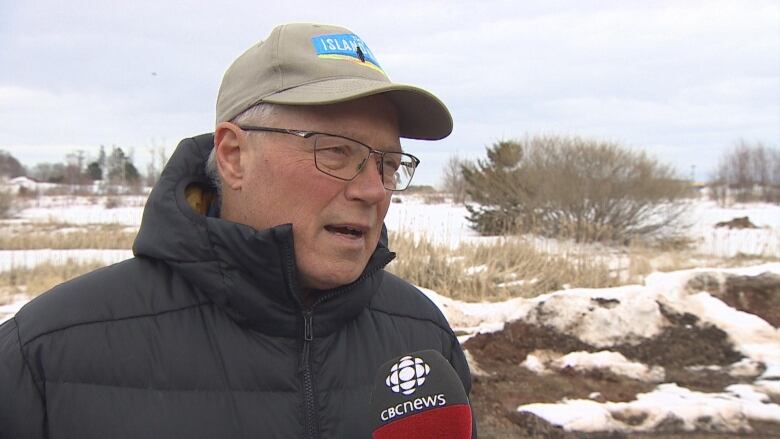 A man in a hat speaking in front of a microphone with the CBC News logo.