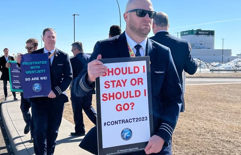 A bald man wearing sunglasses and a suit holds a sign that reads 'should I stay or should I go?.'