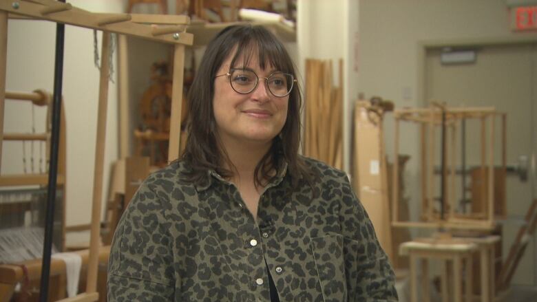 A young woman with glasses sits in a studio with weaving looms in the background.