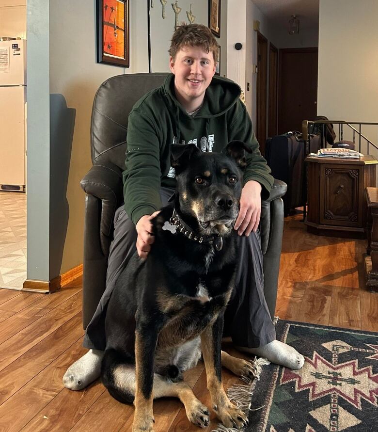 A teenage boy sits on a chair and smiles as he pets his dog.