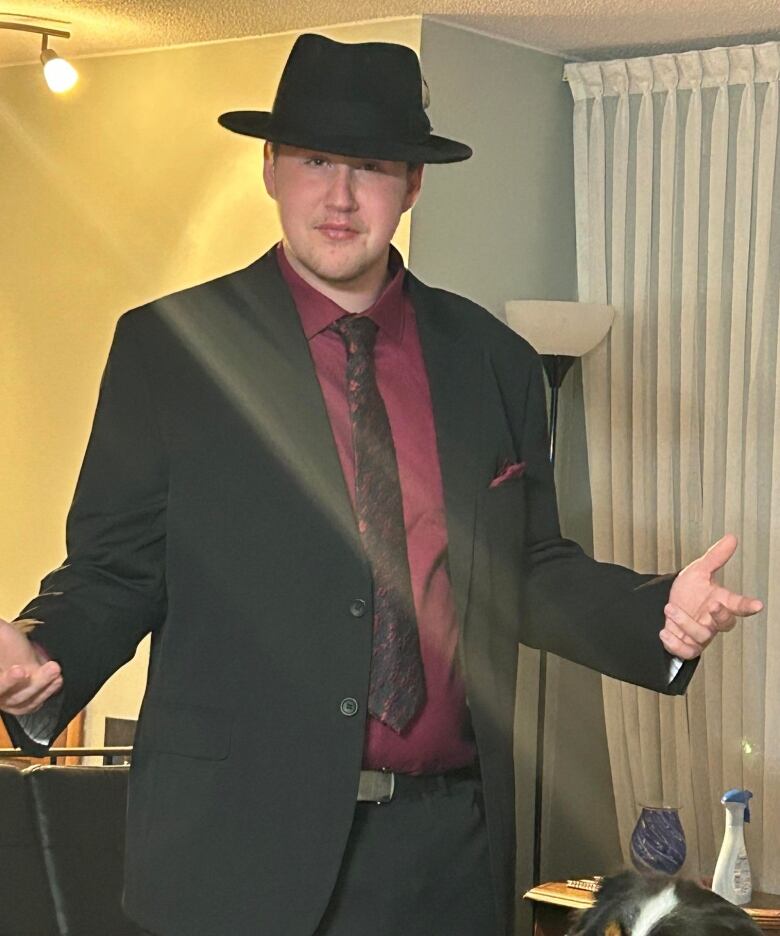 A teen boy wearing a suit and hat smiles and holds his hands up.