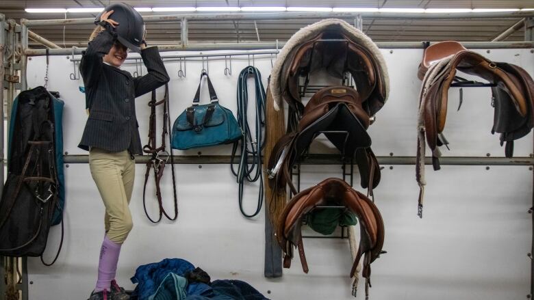 A young girl standing in front of saddles on a wall puts on an English riding helmet.