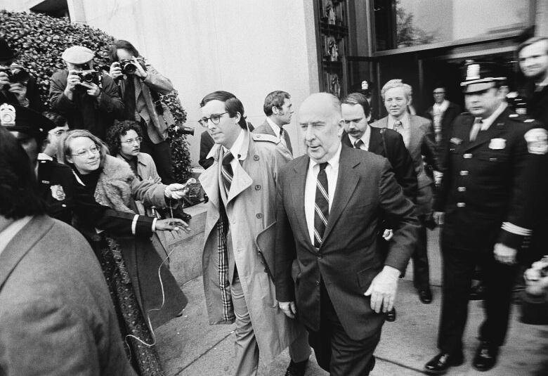 A black and white photograph of two men in suits leaving a building is shown.