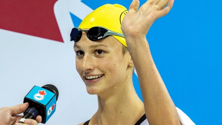 A female swimmer waves with her left hand outside of the pool in front of a hand holding a microphone.