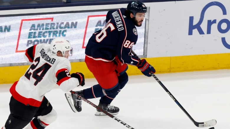 Columbus forward Kirill Marchenko wears a navy blue jersey and skates by Ottawa Senators defenceman, Jacob Bernard-Docker. 