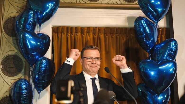 A man in a dark suit and glasses raises both fists in victory on a stage with a microphone. Behind him on stage are heart-shaped blue balloons.