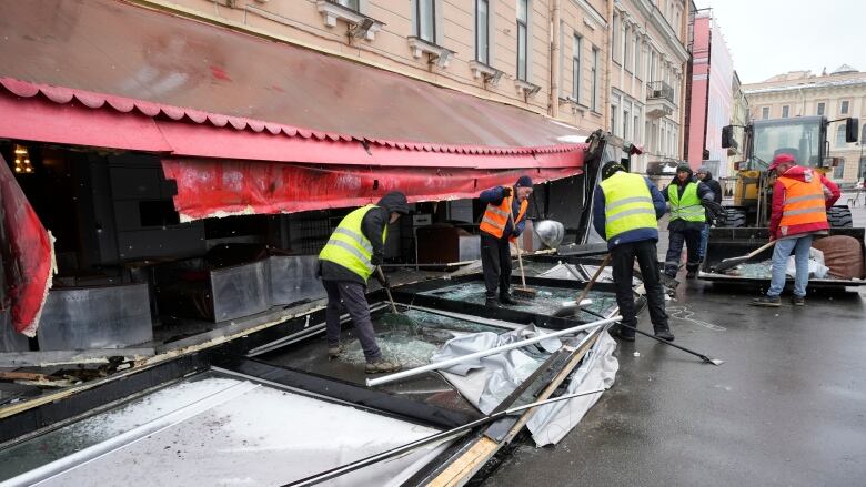 Workers in reflective vests clean up debris outside a building with an awning.