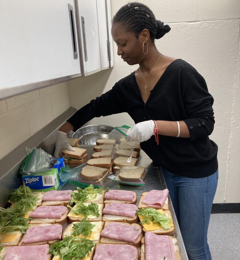 A Black woman is adding lettuce to a long row of ham sandwiches in the cafeteria.