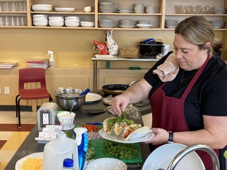 A woman in a cafeteria kitchen adds lettuce to several taco wraps on a plate.