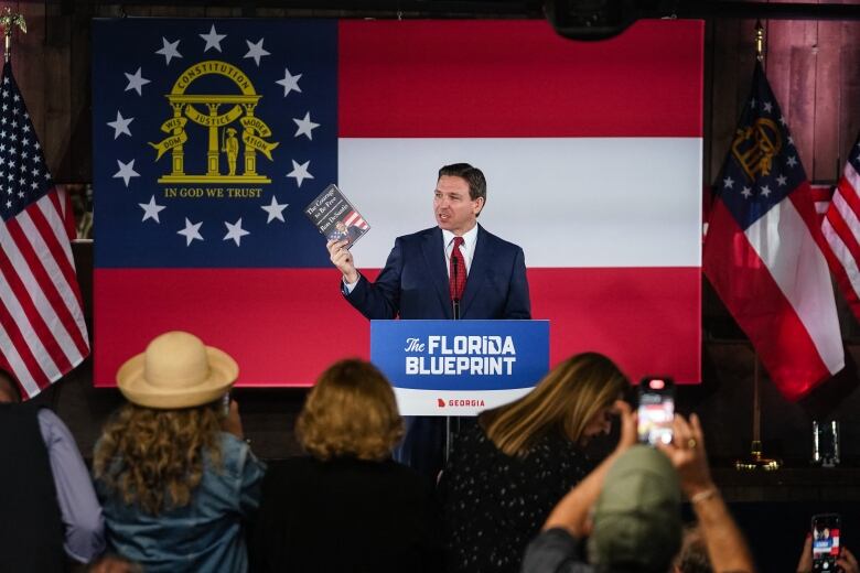 A man in a navy blue suit and red tie stands behind a podium holds up a book in his right hand, with a large flag behind him. 